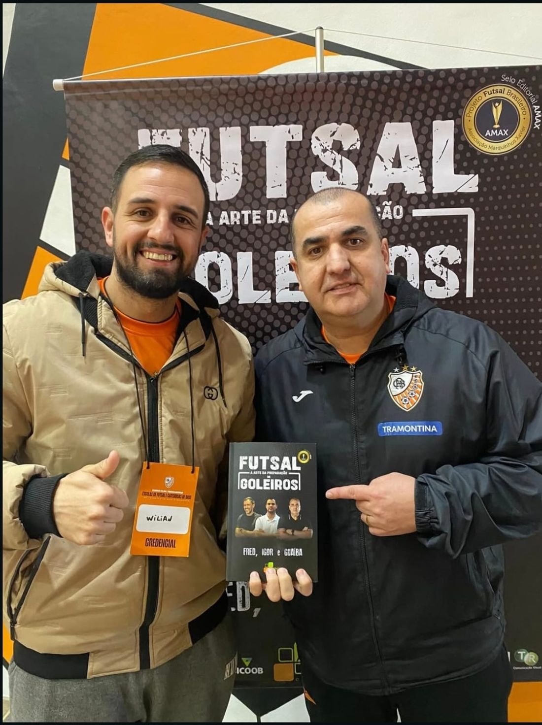 Professor Wilian Silveira Lima, com a credencial do curso, faz sinal de positivo ao lado de um palestrante que segura o livro Futsal: a arte da preparação de goleiros